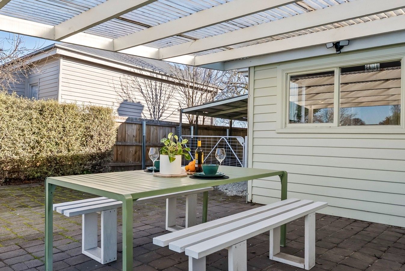 A patio with a table and benches, a potted plant, and a bottle of wine.