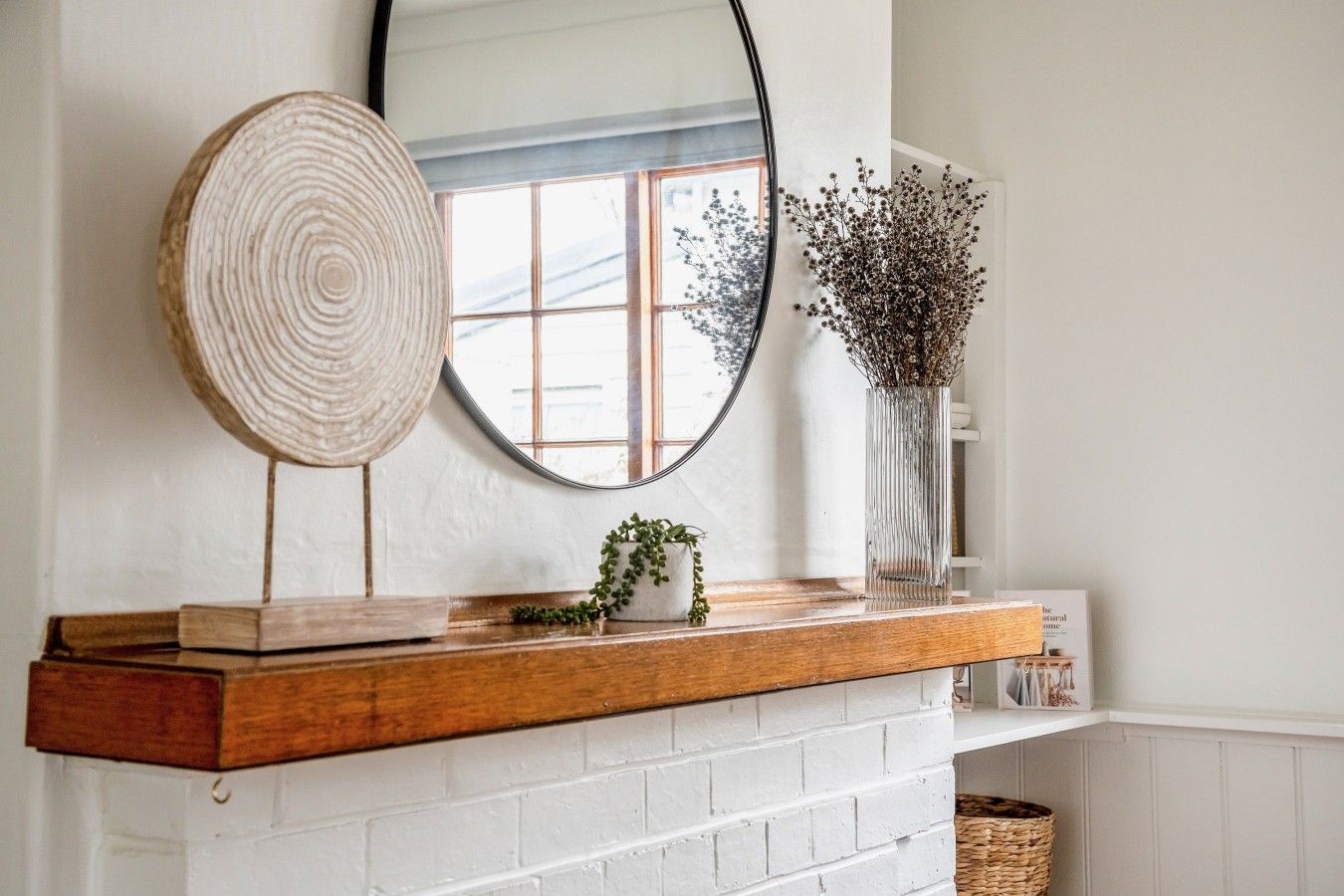 A fireplace mantel with a mirror, a wooden bowl, and a vase with dried flowers.