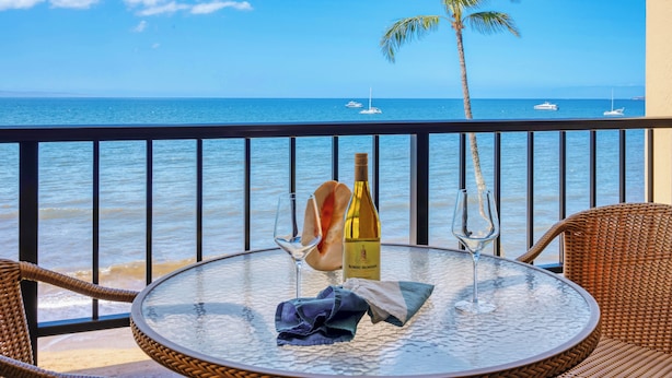Una mesa redonda de vidrio con una botella de vino, una copa y una servilleta en un balcón con vista al océano, barcos y una palmera.