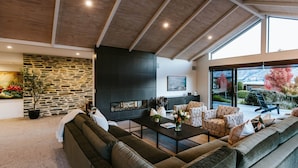 A modern living room with a stone wall, a fireplace, and a large window overlooking a garden. - The Clearview House - Lake Views (Wanaka)