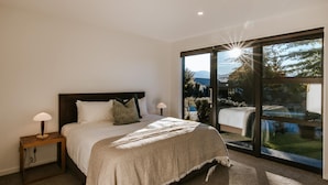 A bedroom with a large bed, two bedside tables with lamps, a view of a garden through a sliding glass door, and a mountain in the background. - The Clearview House - Lake Views (Wanaka)