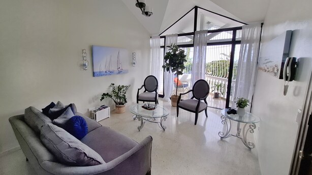 A modern living room with a grey sofa, two armchairs, a glass coffee table, and a potted plant. There's a painting on the wall and a large window with curtains. - Spacious 2BR, Fast WiFi/Ocean & Mountain Views, Security & Convenience (Puerto Plata)