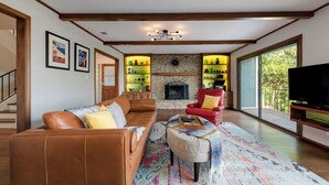 Amplio salón con un sofá de cuero, un sillón rojo y un televisor de pantalla plana grande. En el fondo, una chimenea con detalles de piedra, y estanterías con objetos decorativos a ambos lados.