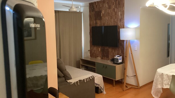 A modern hotel room with a flat-screen TV mounted on a wooden panel wall, a small desk with a lamp, a sofa, and a bed with a white cover.