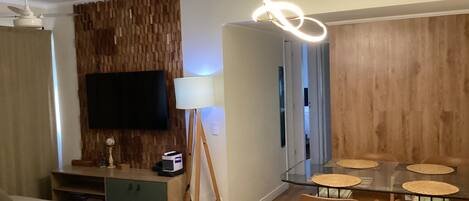 A modern living room with a flat-screen TV, a wooden accent wall, a dining table with chairs, and a floor lamp.