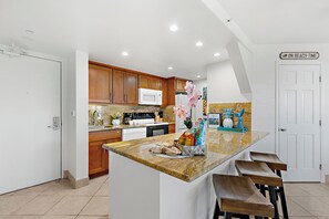 A modern kitchen with wooden cabinets, a granite countertop, and a built-in microwave. There are bar stools and a sign that reads "ON BEACH TIME".