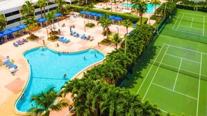 A swimming pool with lounge chairs and palm trees, a tennis court, and a building with multiple floors.