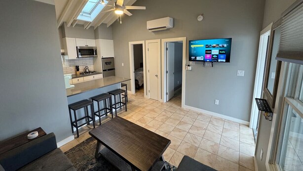Una cocina moderna con microondas integrado, una zona de comedor con mesa de madera y taburetes de bar, y un televisor montado en la pared que muestra servicios de streaming.