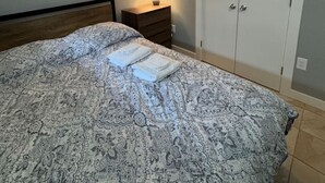 Un dormitori amb un edredó estampat, una làmpada de nit, aire condicionat, un armari i una fotografia emmarcada a la paret.