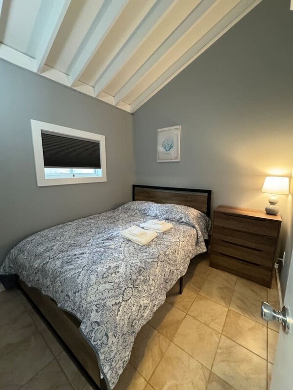 Un dormitorio con cama, mesita de noche, ventana con persianas, un cuadro enmarcado en la pared y una puerta con picaporte.