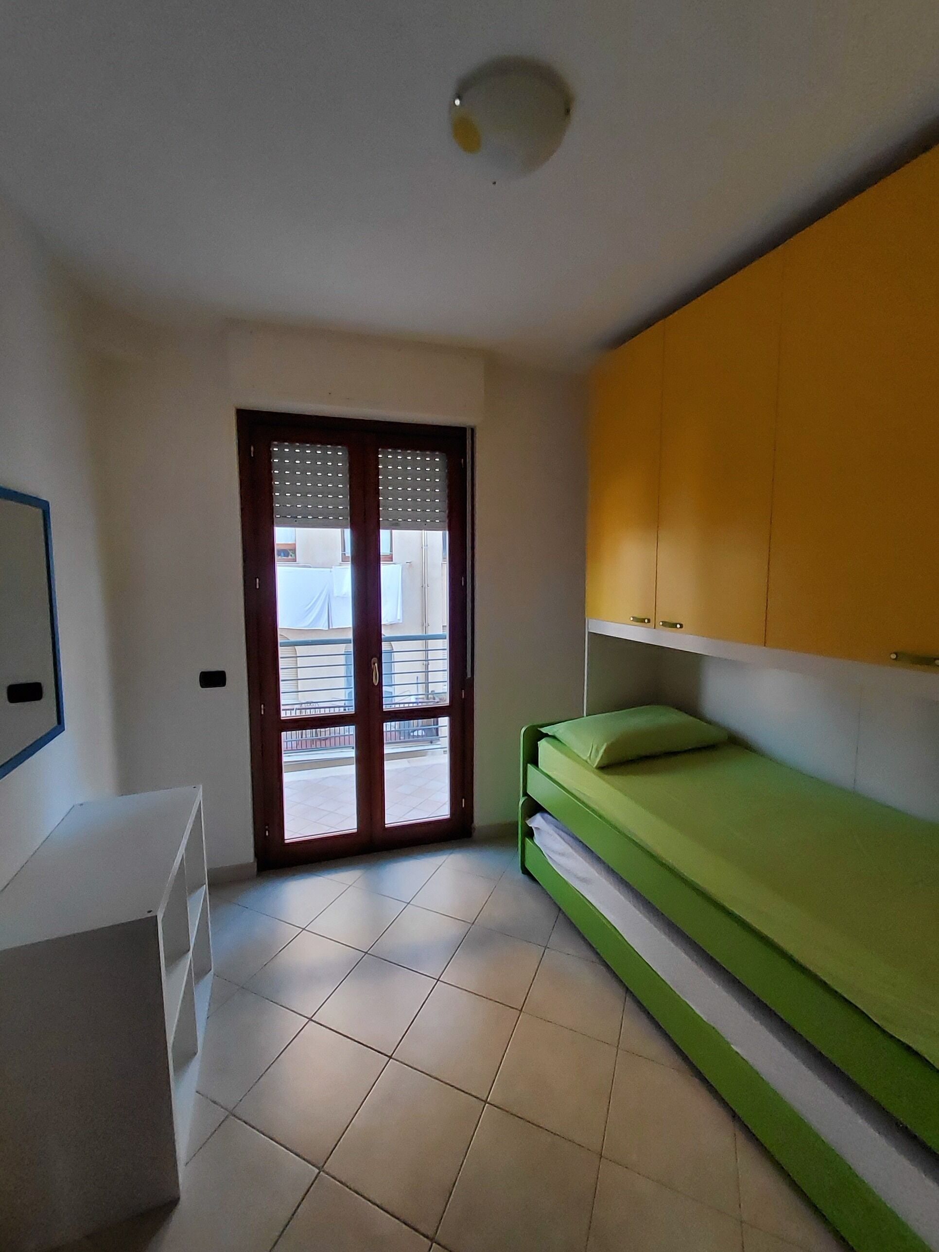 A room with a green bed, a white desk, a yellow cabinet, and a sliding glass door leading to a balcony.