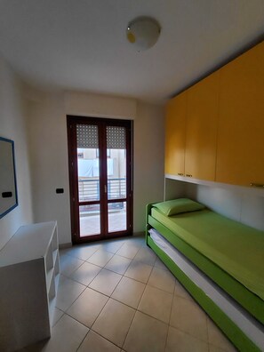 Quarto com cama verde, escrivaninha branca, armário amarelo e porta de vidro deslizante para uma varanda.