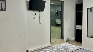 A bedroom with a bed, a flat-screen TV mounted on the wall, a door leading to a bathroom, and a skylight above.
