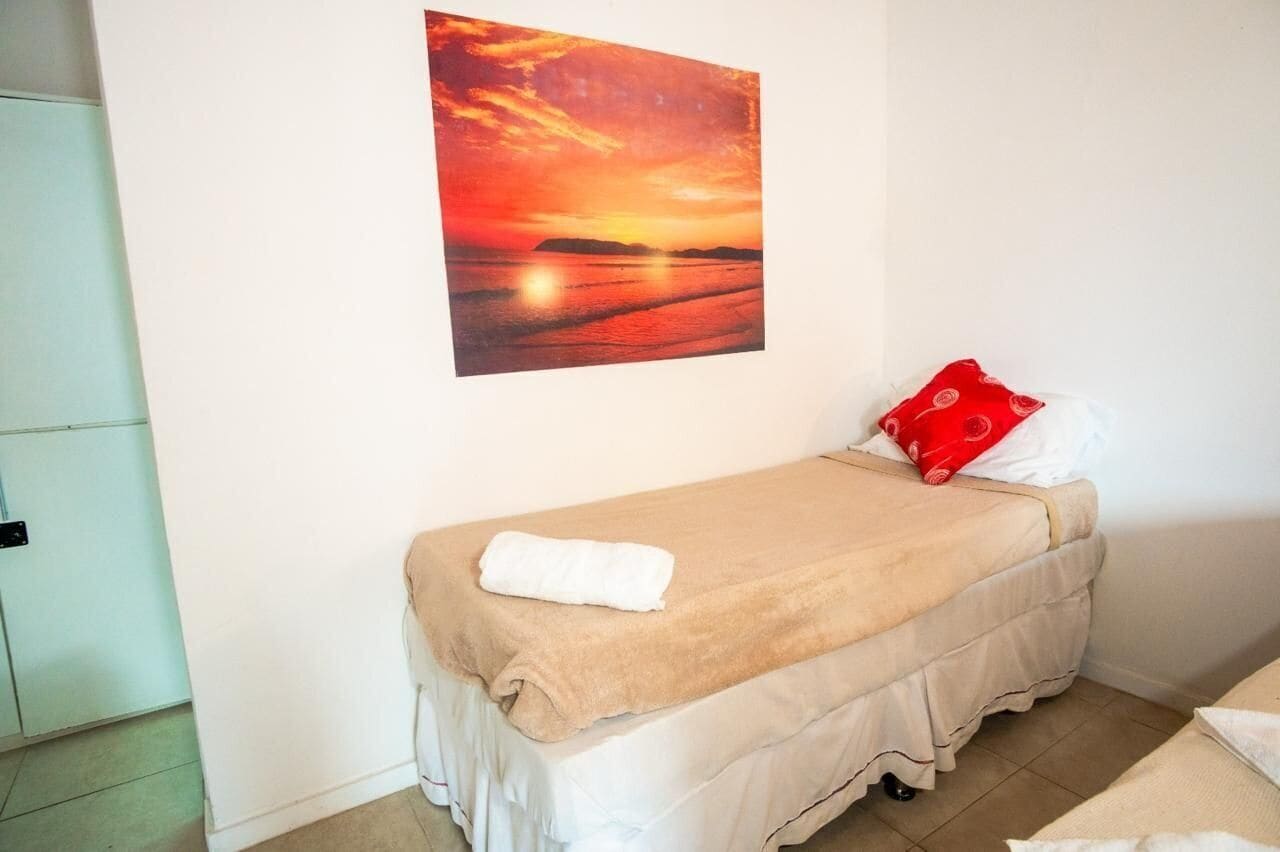 A single bed with a beige bedspread, a white pillow, and a red pillow with a circular pattern. A white towel is folded on the bed.