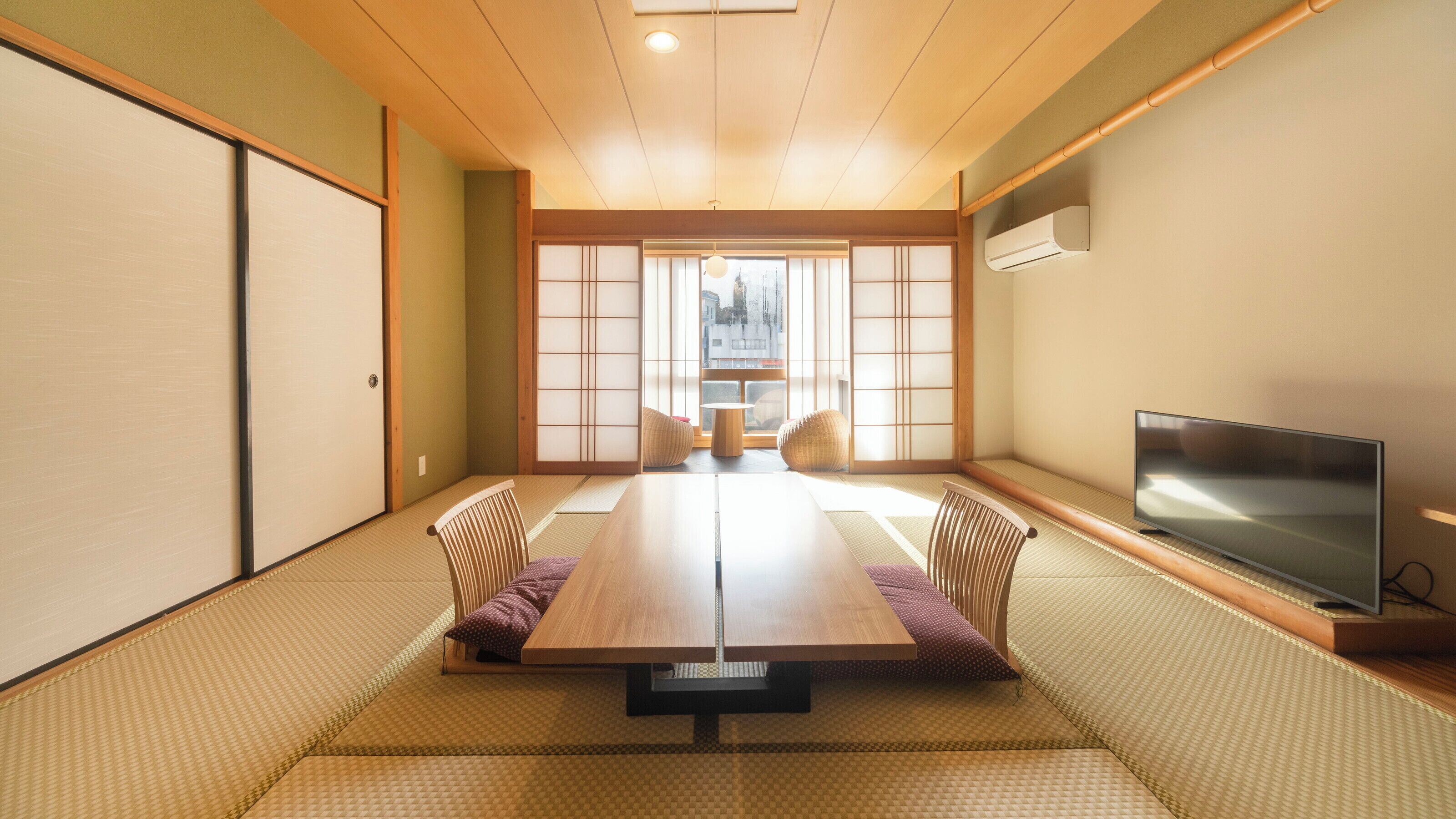 A tatami mat room with a low table, chairs, a television, and a view through a shoji screen.