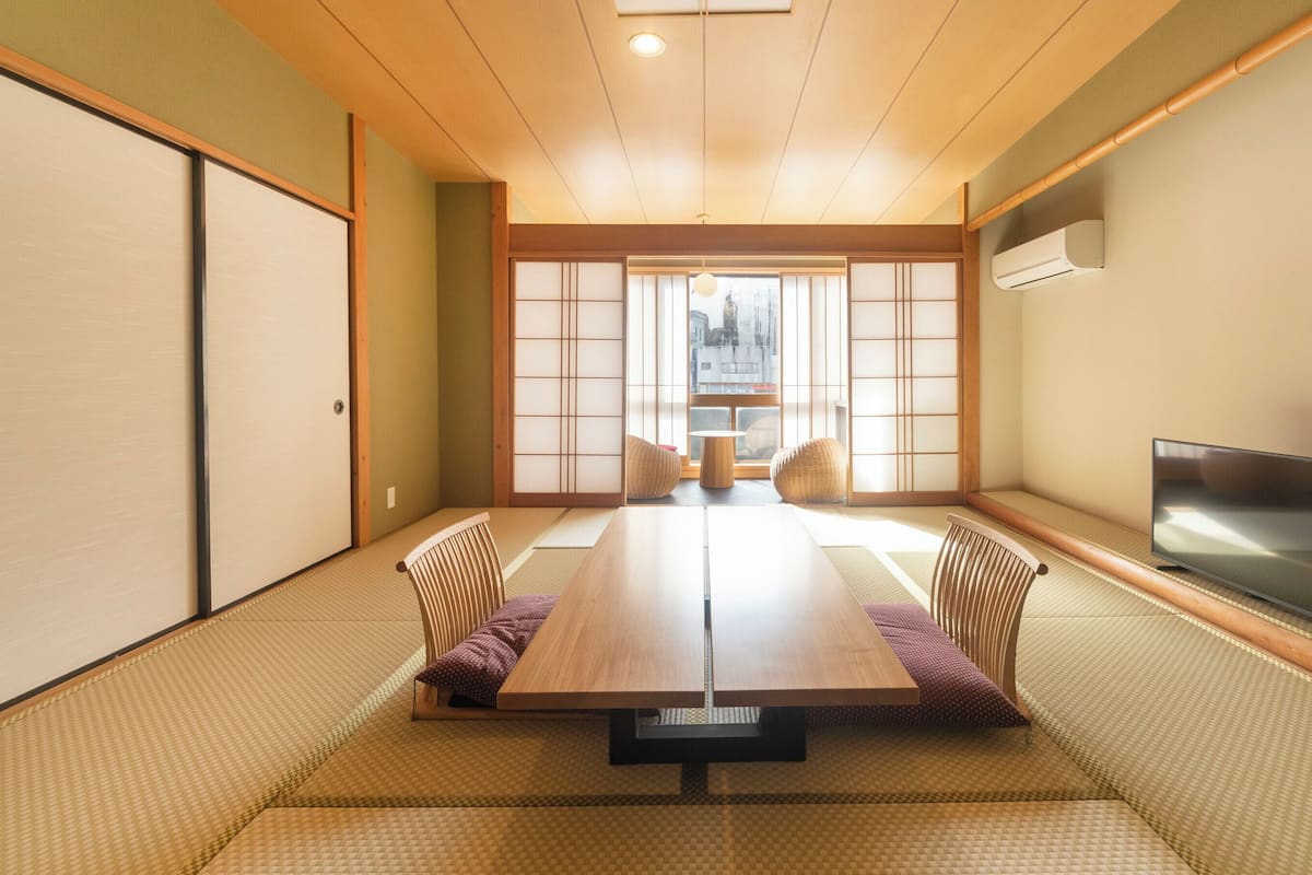 A tatami mat room with a low table, chairs, a television, and a view through a shoji screen.