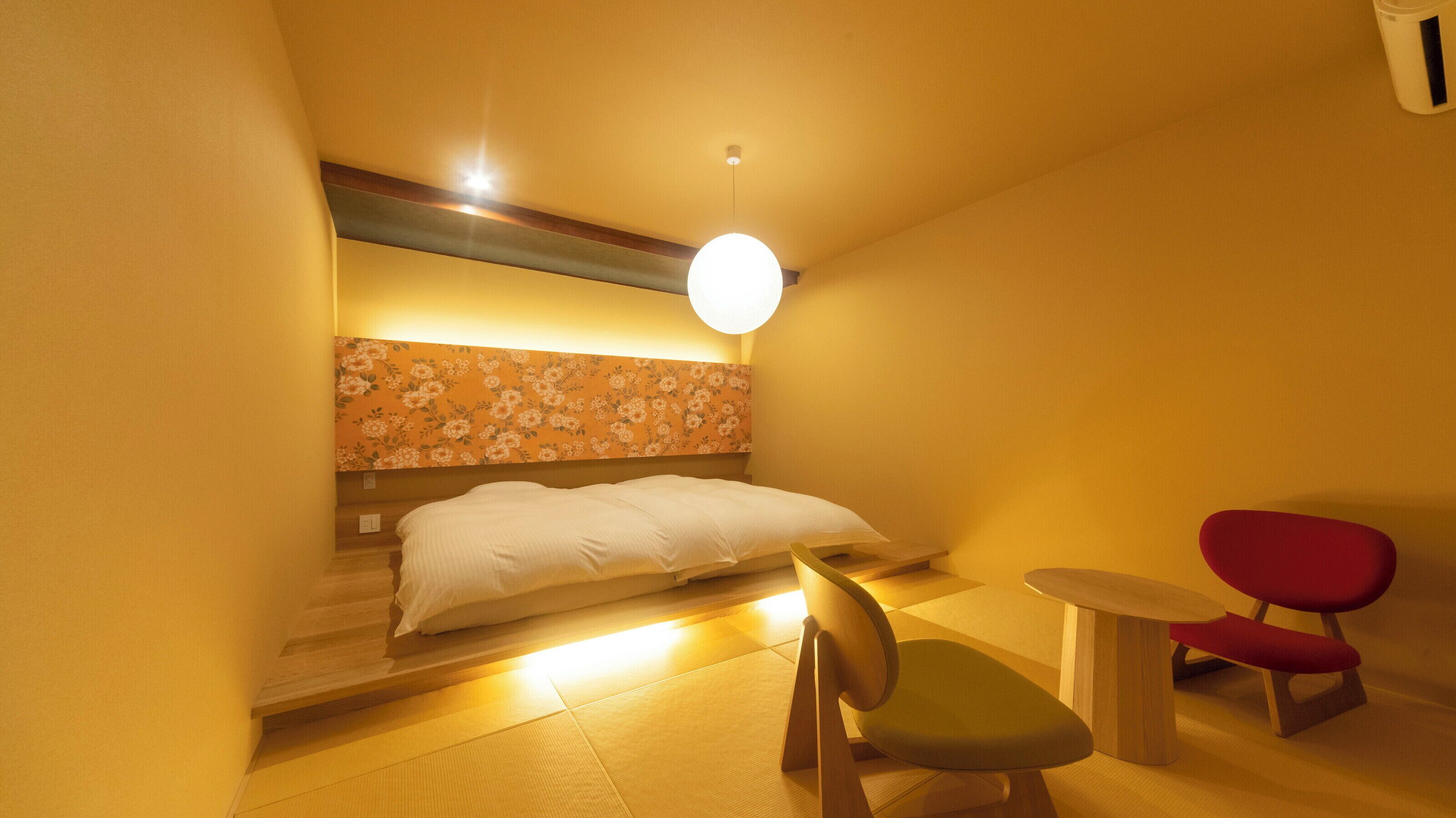 A hotel room with a bed, a chair, a small table, and a wall-mounted light fixture.