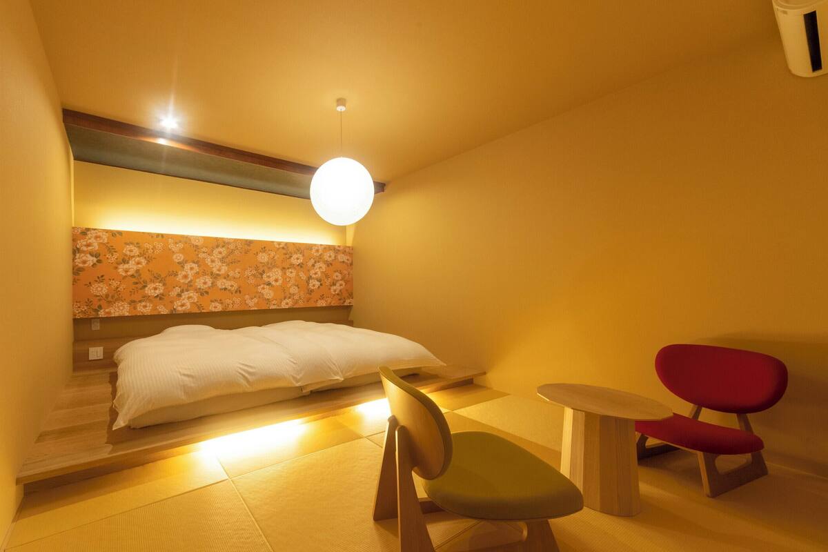 A hotel room with a bed, a chair, a small table, and a wall-mounted light fixture.