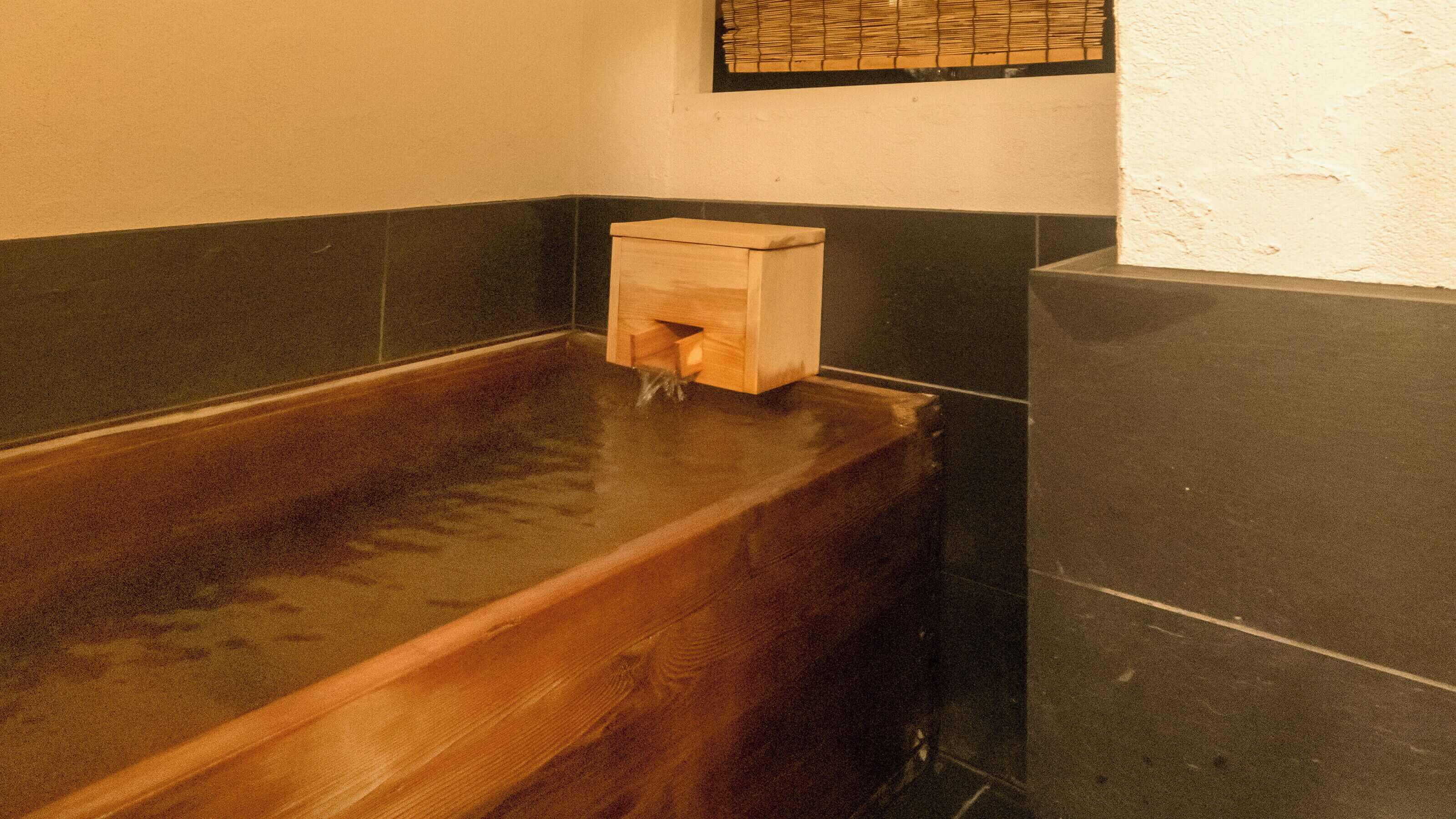 A wooden bathtub with a wooden box in the corner, a tiled wall, and a window with blinds.
