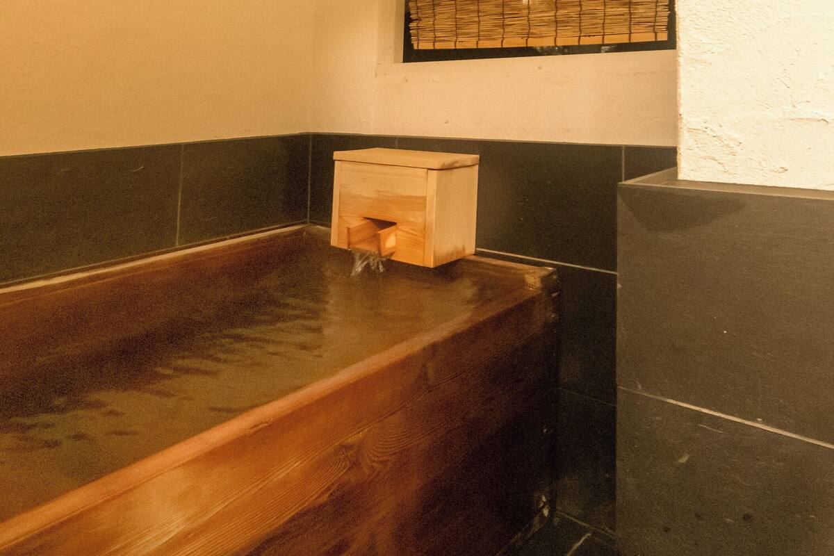A wooden bathtub with a wooden box in the corner, a tiled wall, and a window with blinds.