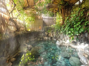 Un área de aguas termales con una piscina revestida de piedra, cerca de setos de madera y abundante vegetación.