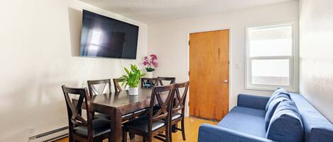 A dining area with a wooden table and chairs, a potted plant, and a flat-screen TV mounted on the wall.