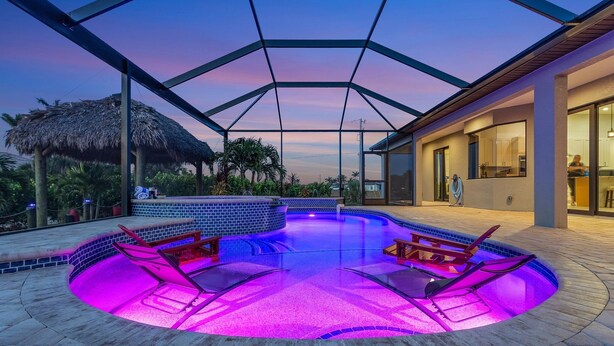 A modern outdoor swimming pool with a glass enclosure, surrounded by a patio area with lounge chairs and a thatched-roof structure.