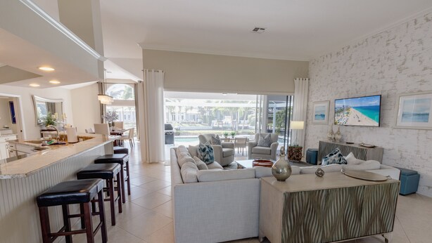 A modern living room with a sofa, coffee table, and bar stools. A large window offers a view of the outdoors. - Waterfront, Private Pool, Walk to Tigertail Beach, Best of Marco! (Marco Island)