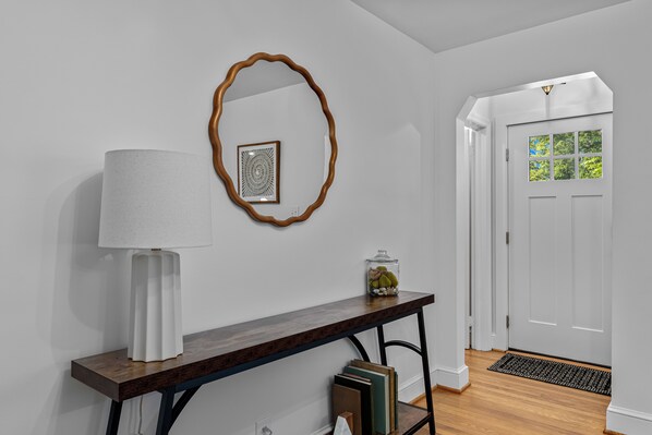 Un pasillo con una consola de madera, una lámpara blanca, un espejo decorativo y una puerta con ventanal.