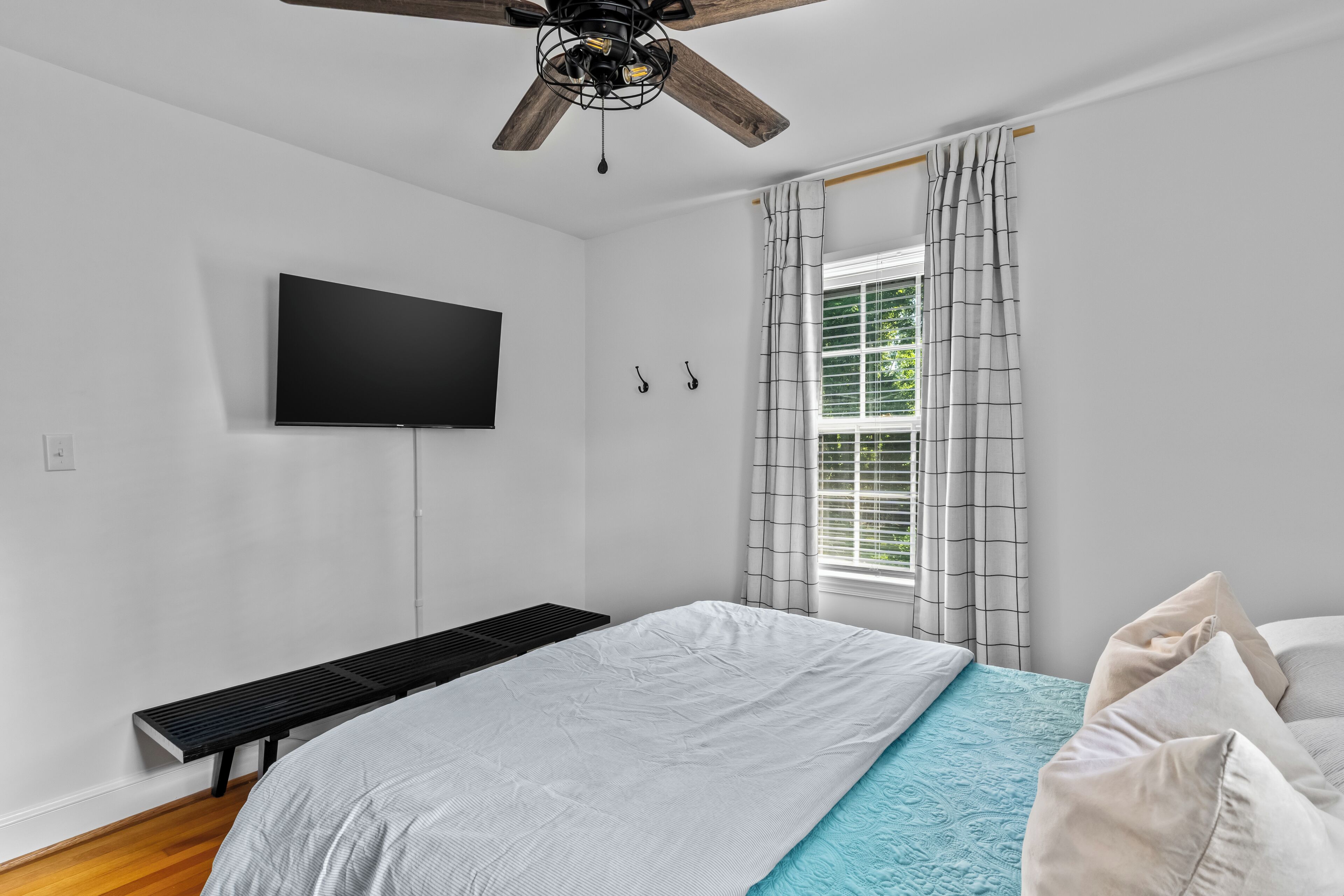 Ein Schlafzimmer mit Bett, Deckenventilator, Wand-Fernseher, Fenster mit Vorhängen und einer Bank.