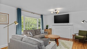 A modern living room with a sectional sofa, a round coffee table, a flat-screen TV mounted above a fireplace, and a green armchair.