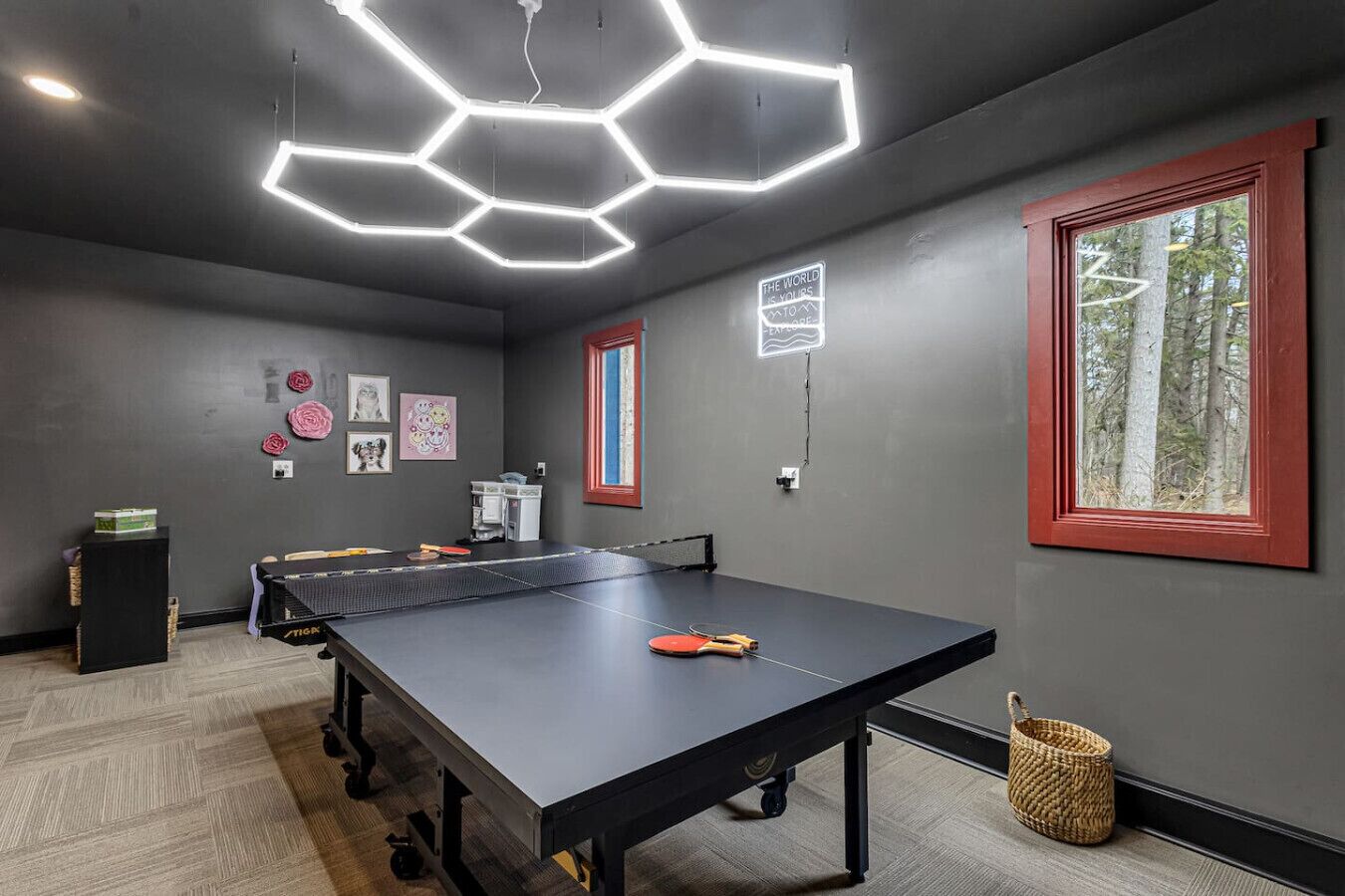 A ping pong table in a room with grey walls, a hexagonal ceiling light, and a window with a red frame.