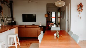 A modern living room with a wooden dining table, white chairs, a leather sofa, a ceiling fan, and a flat-screen TV mounted on the wall. - Evans Head Beach House - Entertainer (Evans Head)