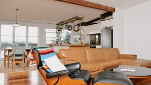 A modern living room with a leather sofa, a dining area with a wooden table and chairs, and a kitchen with hanging pots. - Evans Head Beach House - Entertainer (Evans Head)