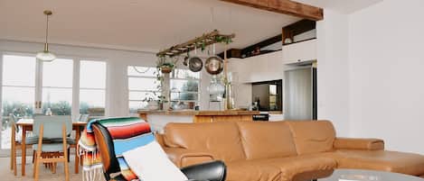 Una sala de estar moderna con un sofá de cuero, una zona de comedor con una mesa y sillas de madera, y una cocina con ollas colgantes.