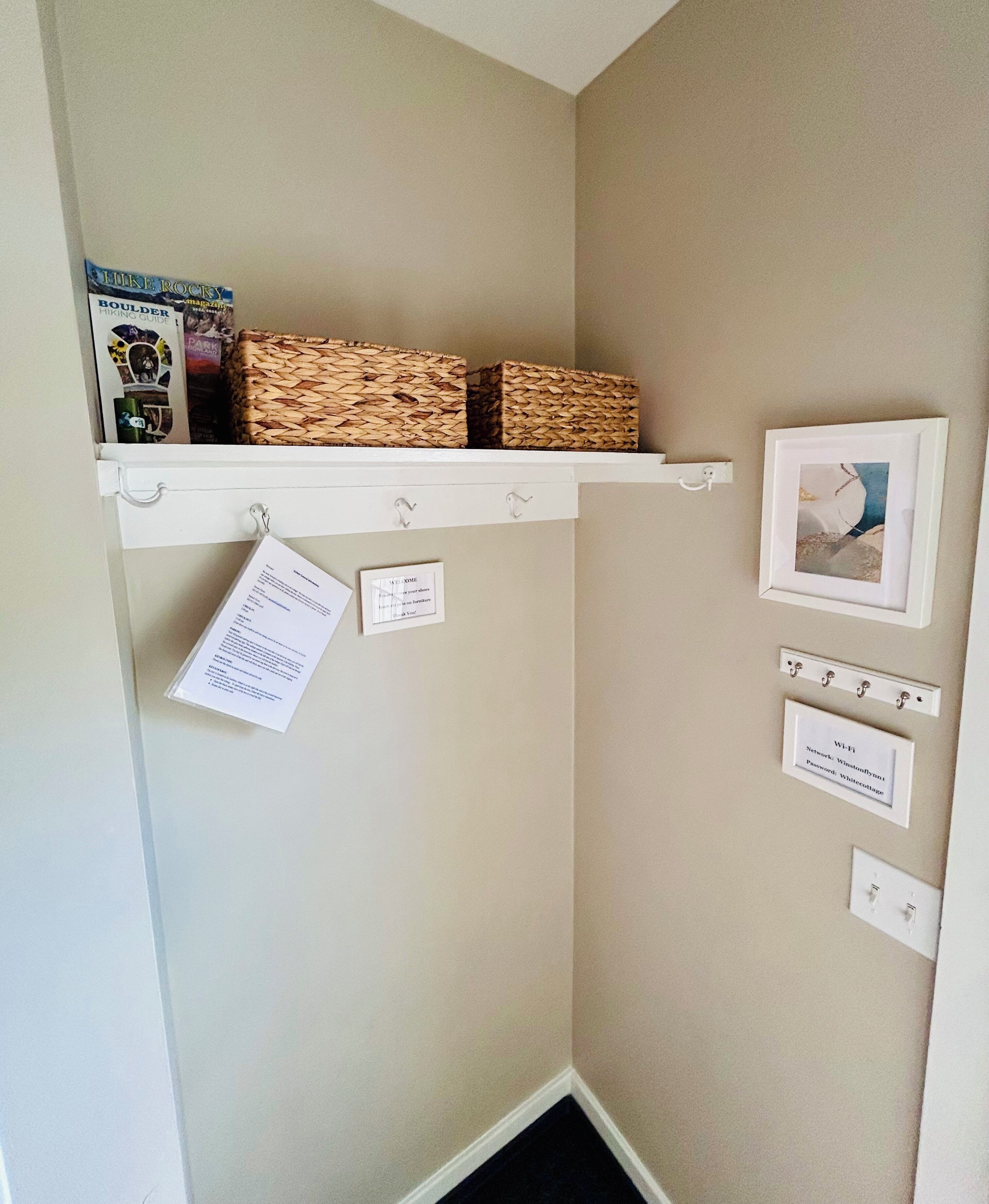 A corner of a room with a white shelf holding a book and two woven baskets. There are two small signs on the wall, one with text and another with a picture.