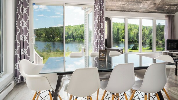 Ein moderner Essbereich mit einem Glastisch und weißen Stühlen, große Fenster mit Blick auf einen See und einen Wald, und eine rustikale Decke.