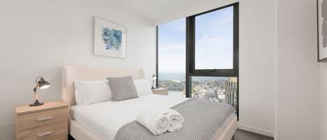 Un dormitorio moderno con una cama grande, una mesita de noche con lámpara, una obra de arte enmarcada en la pared y un amplio ventanal con vistas a la ciudad.