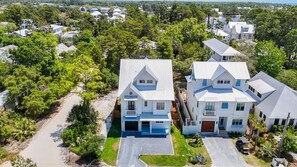 A large, two-story house with a garage, surrounded by lush greenery and other houses. - Shore Thing | Private Pool | Walk to the beach | Bike to Seaside | Brand New Construction and Decor (Santa Rosa Beach)