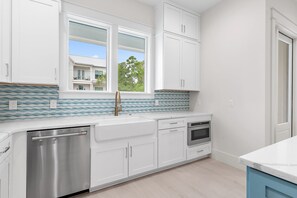 A modern kitchen with white cabinetry, a stainless steel dishwasher, an oven, and a window with a view of a house and trees. - Shore Thing | Private Pool | Walk to the beach | Bike to Seaside | Brand New Construction and Decor (Santa Rosa Beach)