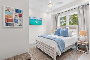 A bedroom with a bed, a nightstand, a lamp, a wall-mounted TV, and a framed collection of vintage-style postcards. - Shore Thing | Private Pool | Walk to the beach | Bike to Seaside | Brand New Construction and Decor (Santa Rosa Beach)