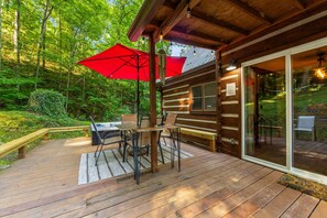 Une terrasse en bois avec une table et des chaises, un parasol rouge et une porte vitrée donnant sur un jardin.