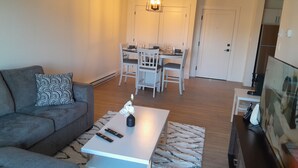 Una sala de estar con un sofá gris, una mesa de centro con controles remotos, un cojín estampado, una mesita con un florero, y una zona de comedor con una mesa y sillas blancas.