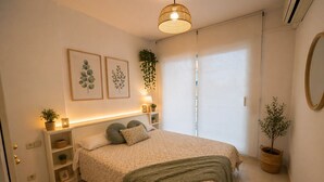 Un dormitorio con cama, mesitas de noche, una planta colgante, una lámpara de pared y obras de arte enmarcadas.