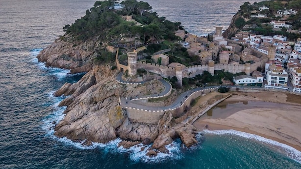 Vista aérea de un pueblo costero con una estructura fortificada, una playa y un litoral rocoso.