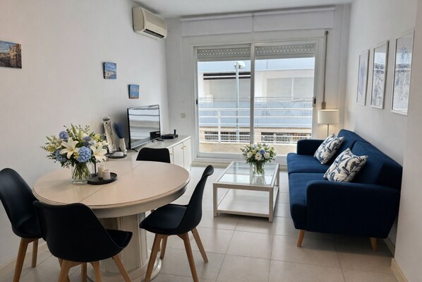 A modern living room with a round dining table, two black chairs, a blue sofa, and a balcony with a view.