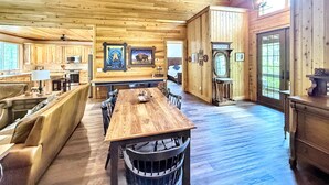 Ruim houten interieur van een chalet met een grote eettafel, gezellige zithoek, een keuken en een groot raam met uitzicht op de natuur.