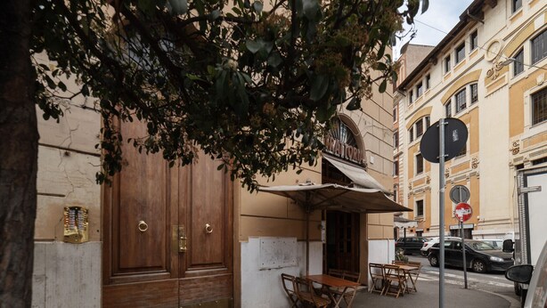 Vista de la calle con un edificio que tiene una puerta de madera grande, un espacio al aire libre cubierto con mesas y sillas, y un letrero con un emblema circular.