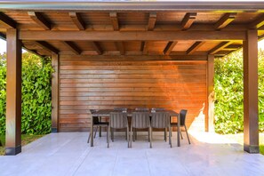 Un patio al aire libre con una pérgola de madera, una mesa rectangular con sillas y piso de baldosas.