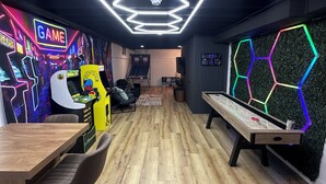 A room with hexagonal ceiling lights, a wooden floor, a shuffleboard table, and arcade machines. - Game Room! Modern 3BR 2.5BA New Build Luxury—Near Downtown Cleveland! (Cleveland)